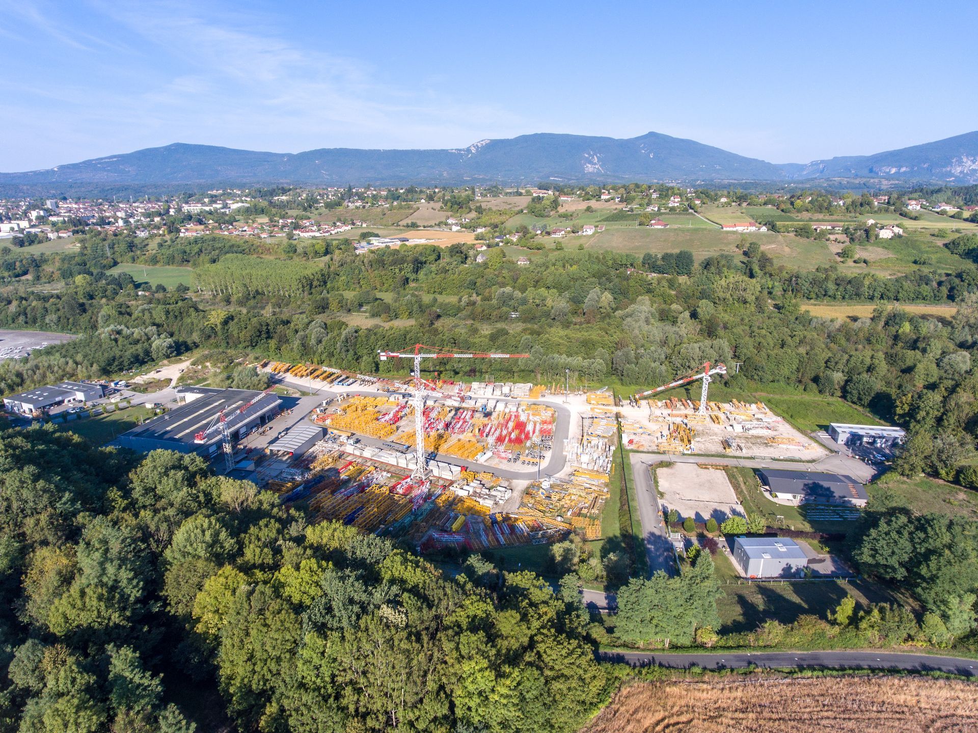 Vue aérienne d'un chantier de construction avec des grues et des matériaux colorés, entouré d'arbres et d'un paysage rural.