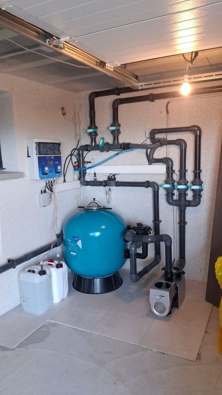 Local technique avec réservoir sous pression bleu, tuyauterie et équipement de pompage sur sol carrelé.