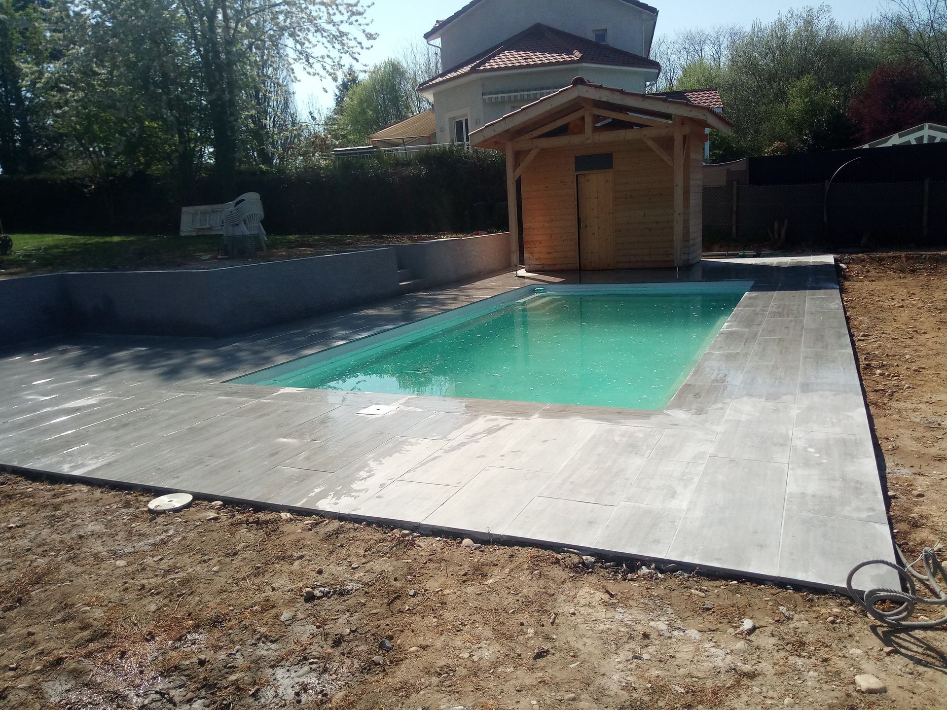 Piscine dans le jardin avec eau turquoise, cabane en bois et terrasse pavée.