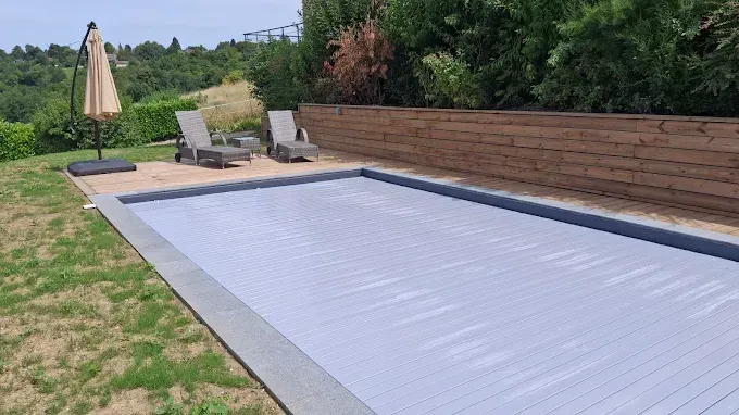Piscine couverte avec chaises longues, parasol beige et clôture en bois dans un jardin.