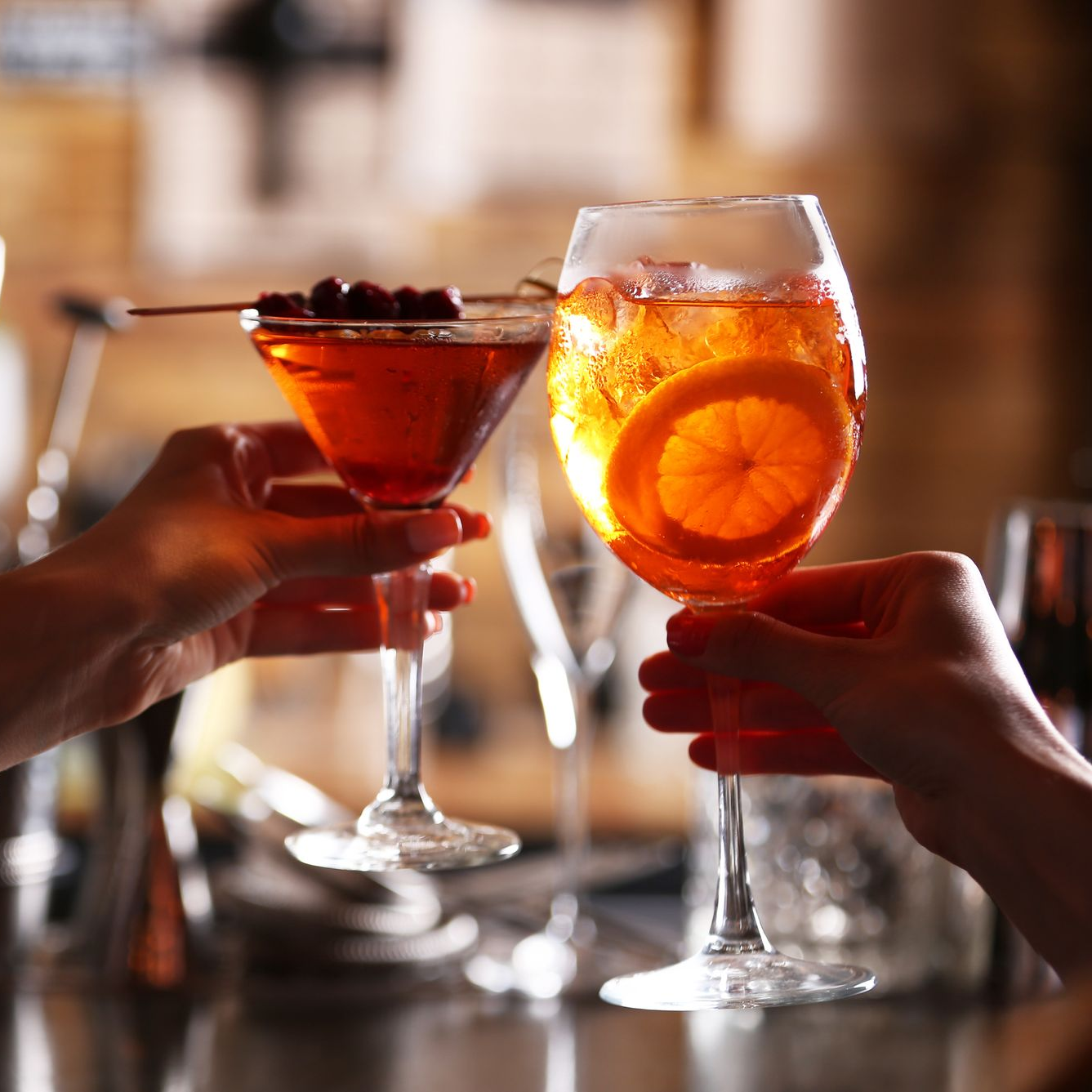 Des mains trinquent avec des cocktails dans un bar à l'éclairage chaleureux : un cocktail à l'orange et un martini rouge aux fruits rouges.