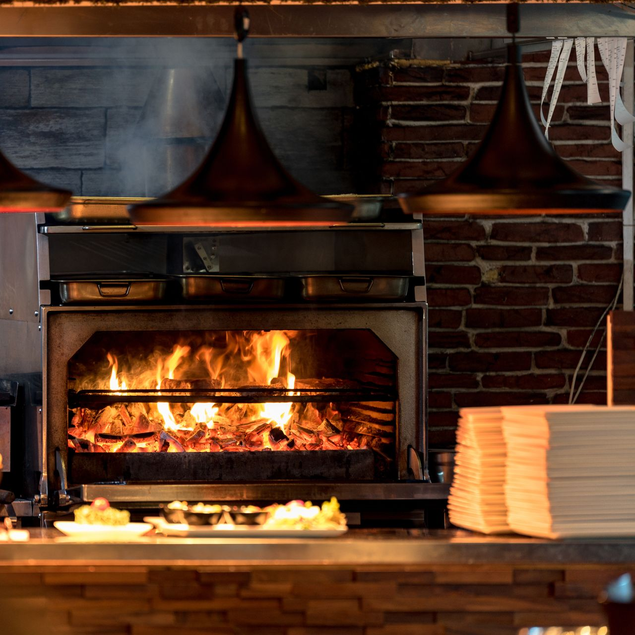 Four à pizza au feu de bois avec des flammes dans une cuisine en briques, assiettes et plateaux empilés à proximité.