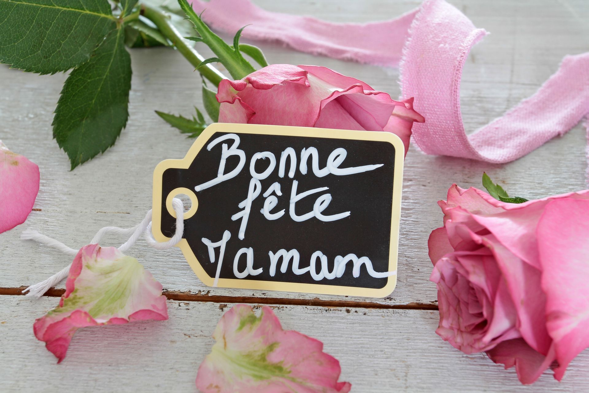 Panneau avec écrit Bonne fête Maman sur un tableau noir avec des roses roses et des pétales sur une surface en bois blanc.