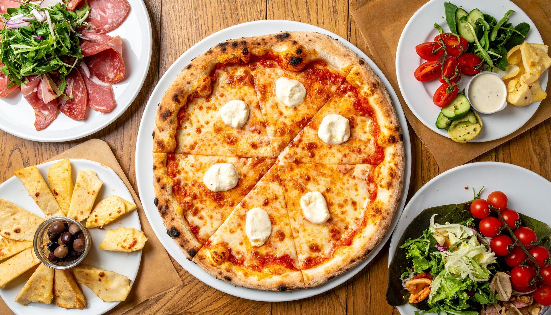 Repas italien copieux avec une pizza au centre, entourée de salades, de charcuterie, de fromages et d'amuse-gueules.