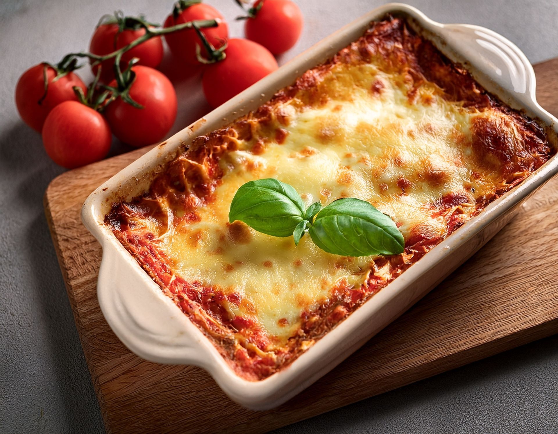 Lasagnes cuites au four dans un plat blanc, gratinées au fromage fondu et au basilic, accompagnées de tomates fraîches sur une planche en bois.