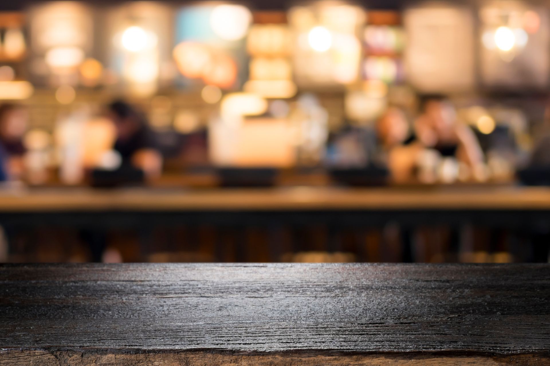 Table en bois dans un café à l'éclairage tamisé, avec des clients flous et des lumières chaudes en arrière-plan.