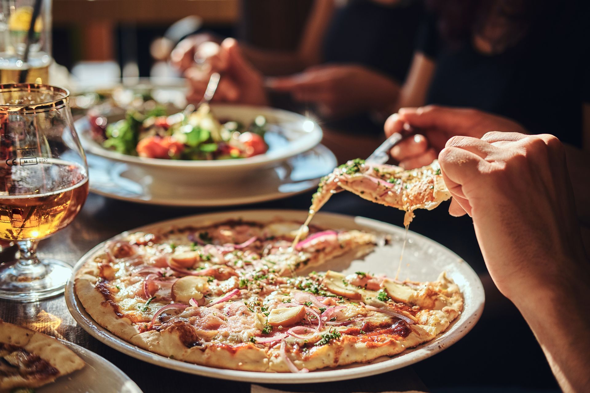 Des mains se partagent une pizza à la table d'un restaurant, accompagnée de salade et de boissons.