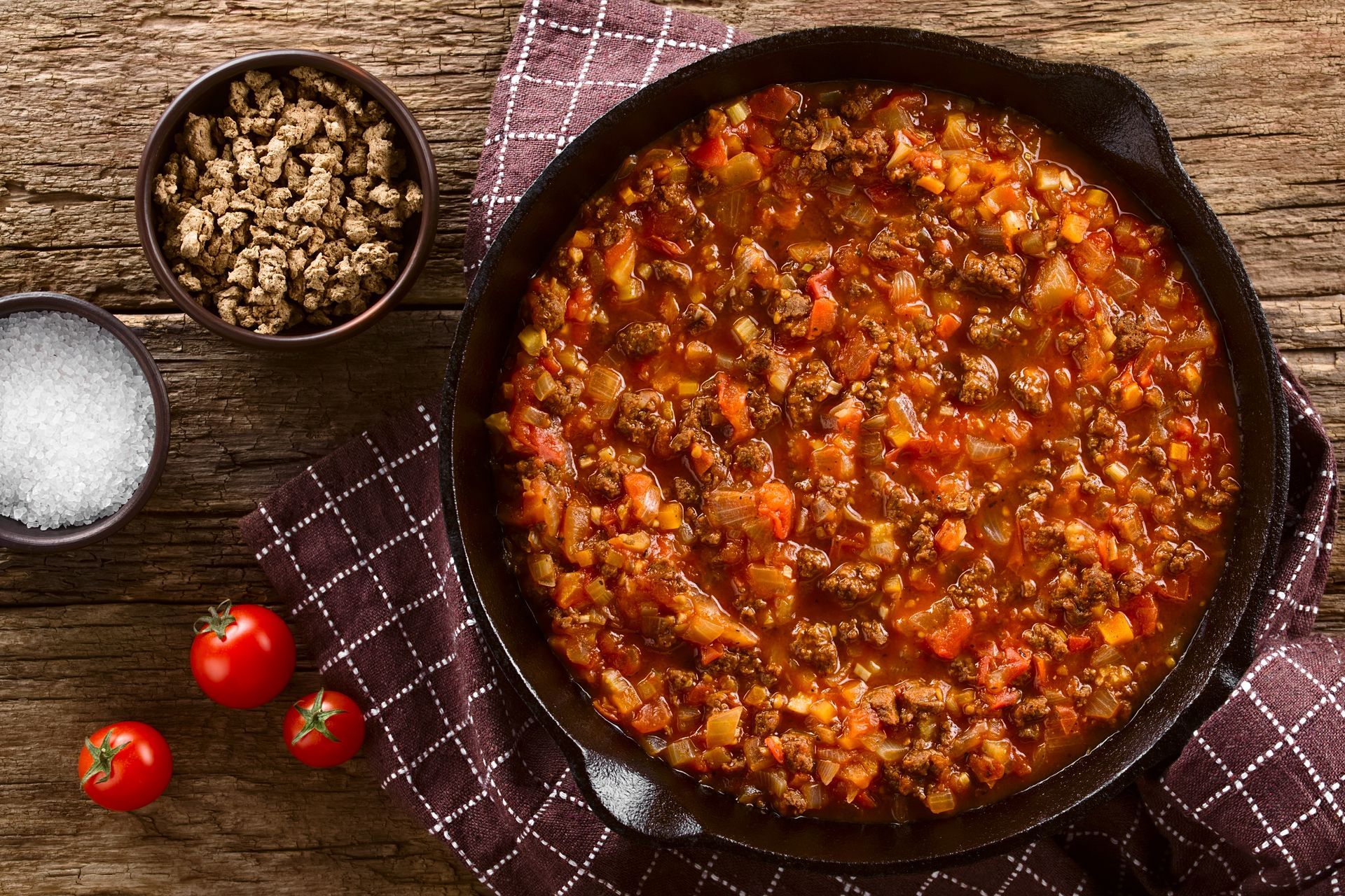 Poêle de sauce tomate épaisse avec un petit bol de fromage émietté, de sel et de tomates cerises sur une table.