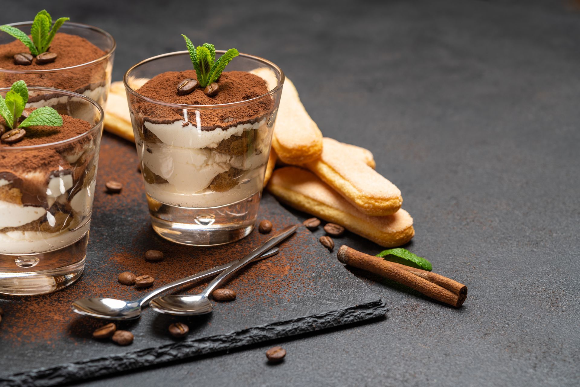 Coupes de dessert avec couches de crème et de cacao sur un fond foncé, garnies de menthe, de cuillères, de gousses de vanille et de biscuits.