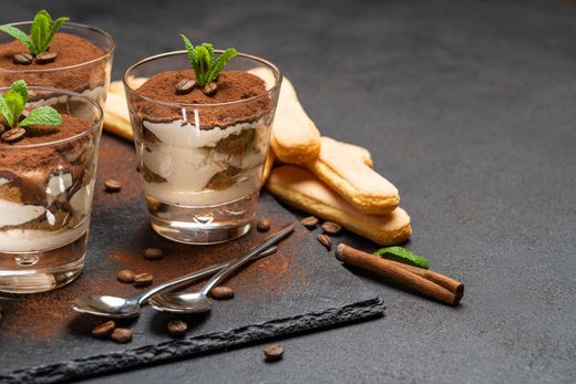 Coupes de dessert avec couches de crème et de cacao sur un fond foncé, garnies de menthe, de cuillères, de gousses de vanille et de biscuits.