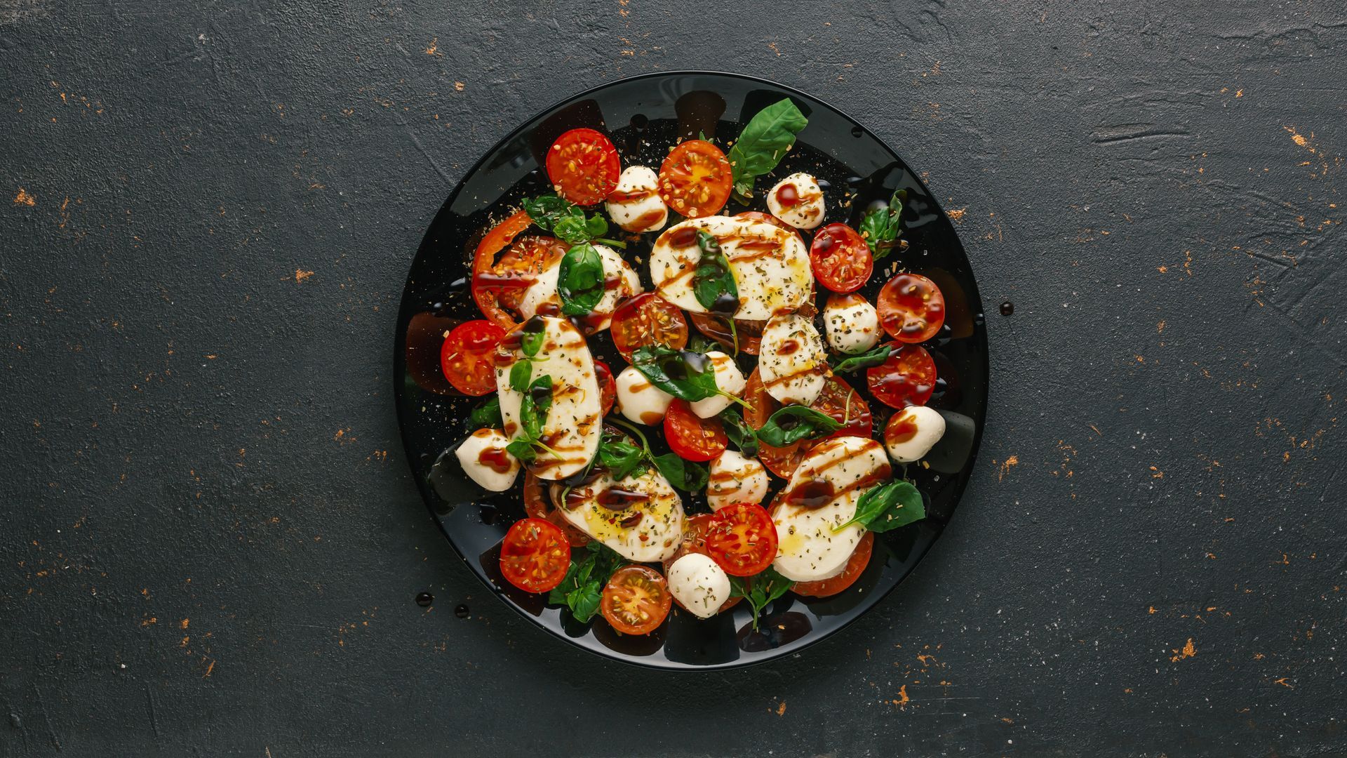 Vue de dessus d'une salade colorée composée de tomates, de mozzarella et de verdure, servie sur une assiette sombre.