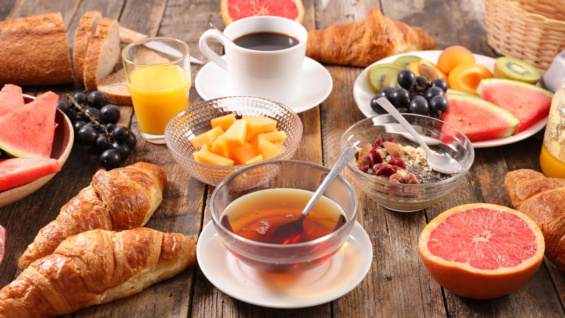 Petit-déjeuner composé de croissants, de fruits, de jus, de café et de confiture sur une table rustique en bois.