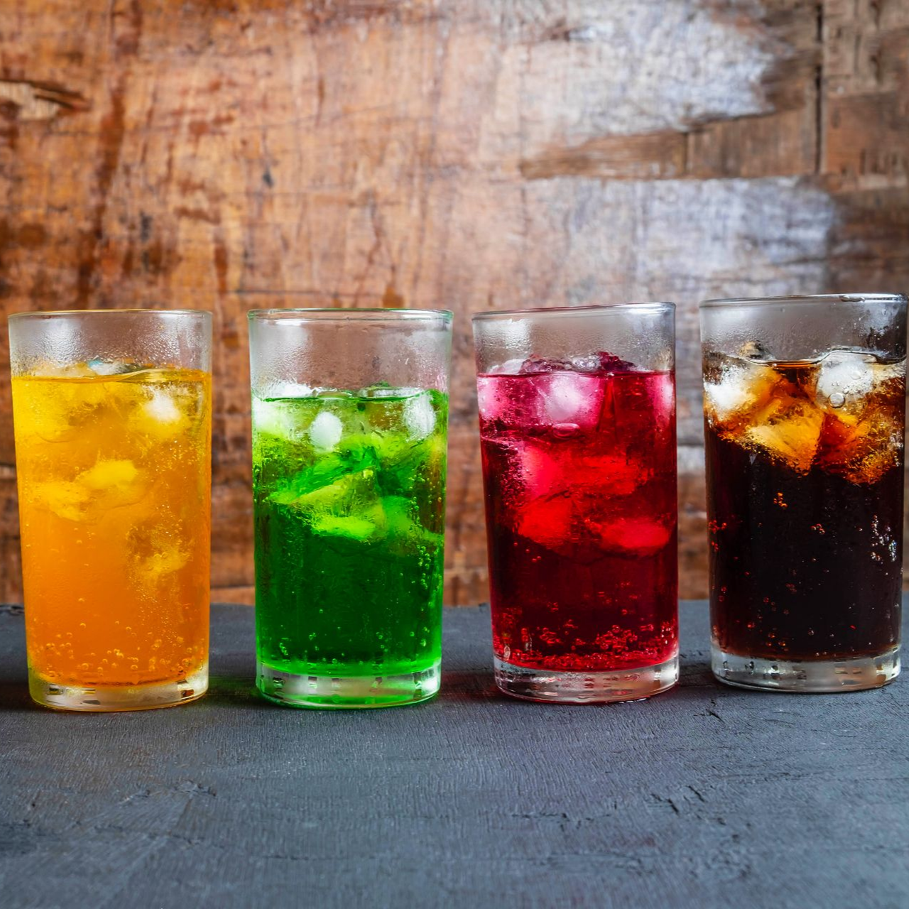 Quatre boissons glacées colorées — orange, verte, rouge et cola noir — alignées sur une table.