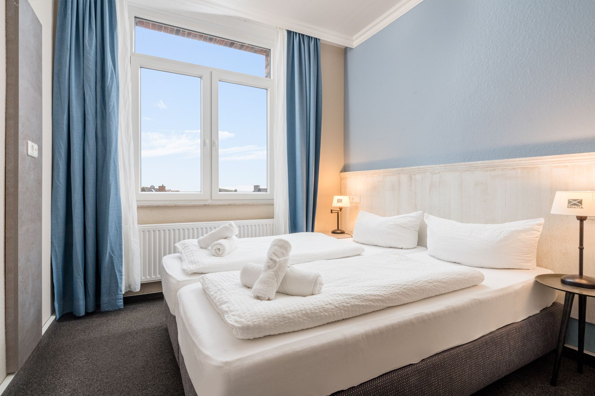 Schlafzimmer mit zwei Betten, weißer Bettwäsche, blauen Vorhängen und hellblauen Wänden.