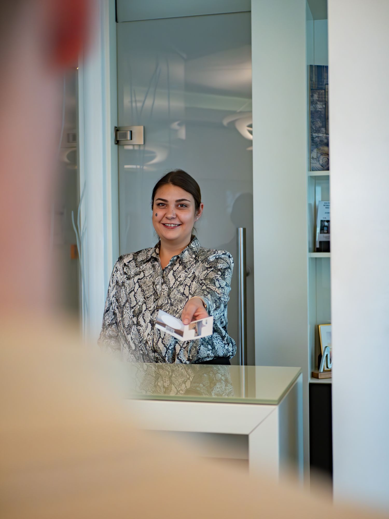 Eine Person in einem gemusterten Hemd lächelt, während sie in einem modernen Büro über den Empfangstresen eine Visitenkarte reicht.