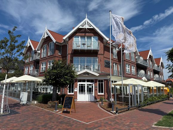 Schlafzimmer mit Bett, Nachttisch und Stuhl. Neutrale Farben mit hellem Holzboden und weißen Vorhängen.