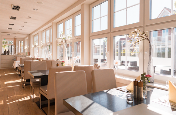 Schlafzimmer mit Bett, Nachttisch und Stuhl. Neutrale Farben mit hellem Holzboden und weißen Vorhängen.