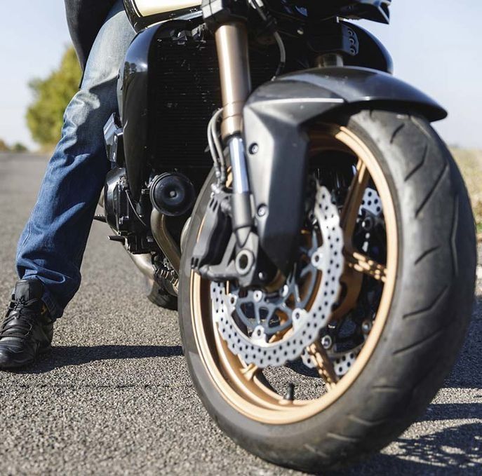 Motocicleta en una carretera asfaltada con la rueda delantera en foco; las piernas del conductor visibles en jeans azules.