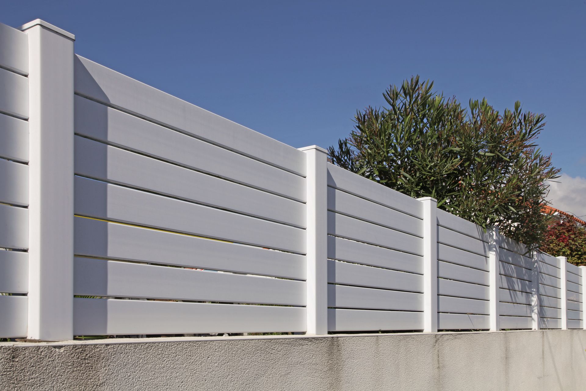 Clôture à claire-voie blanche avec des poteaux gris le long d'un mur de gravier sous un ciel bleu.