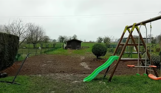 Aire de jeux dans la cour arrière avec un toboggan vert, une balançoire en bois et une pelouse sous un ciel couvert.