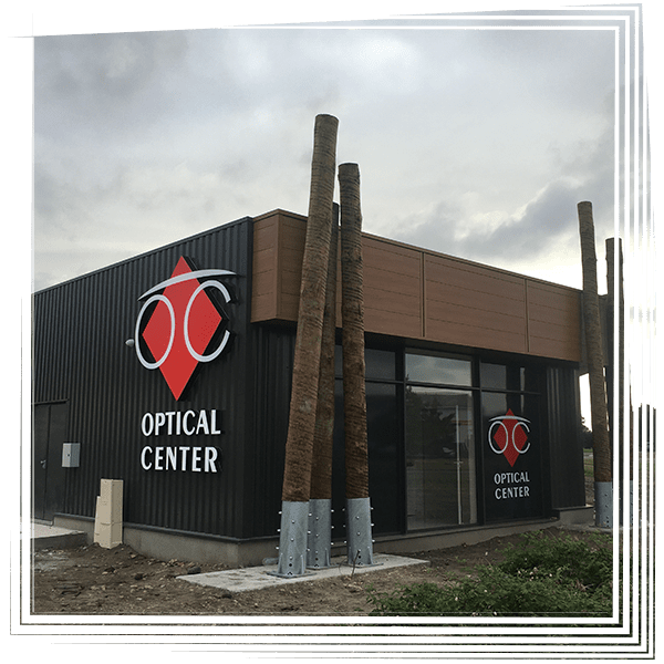 Façade d'un centre d'optique avec logo rouge par temps nuageux, visible à côté de hauts poteaux en bois.