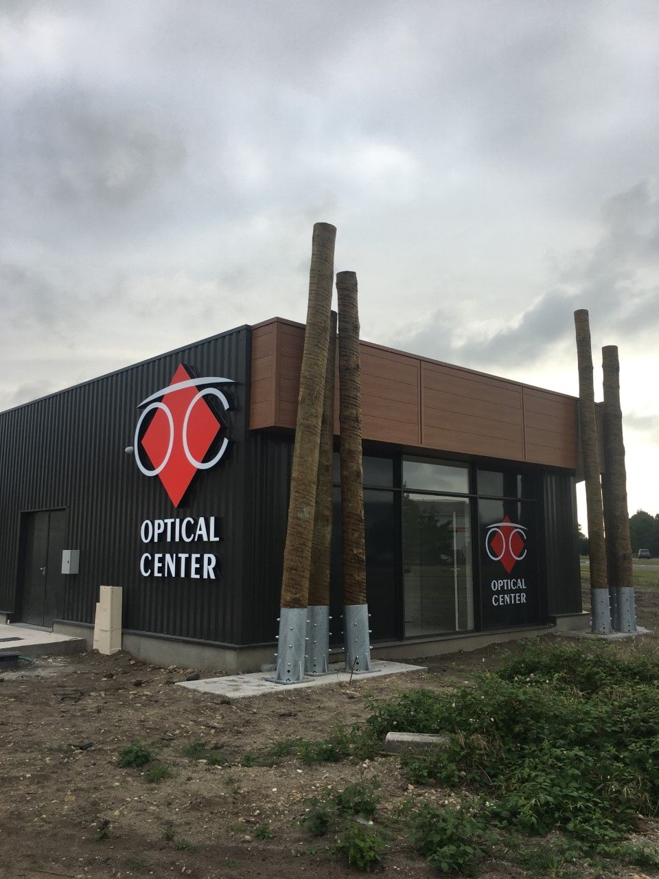 Façade d'un centre d'optique avec logo rouge, façade noire et marron, et ciel nuageux
