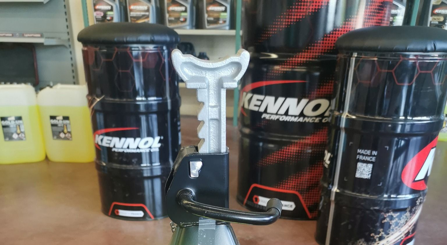 Cric de voiture posé devant des fûts noirs Kennol servant de tabourets. Des bidons jaunes en arrière-plan.