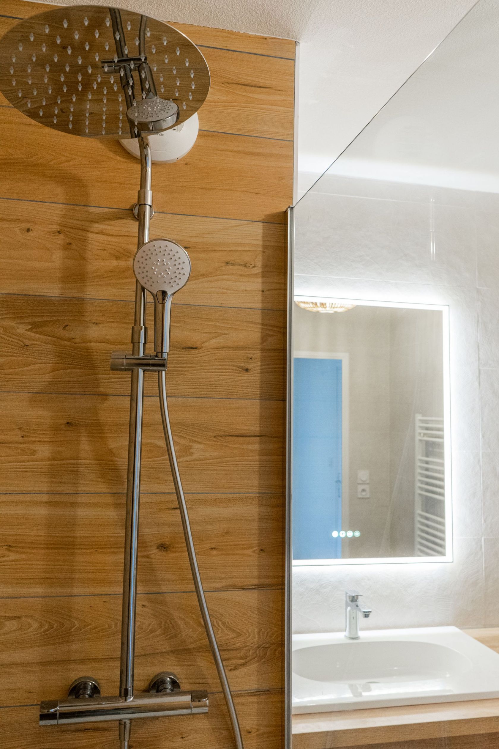 Douche avec paroi en bois et grand pommeau rond. Le miroir reflète un lavabo et une porte bleue.