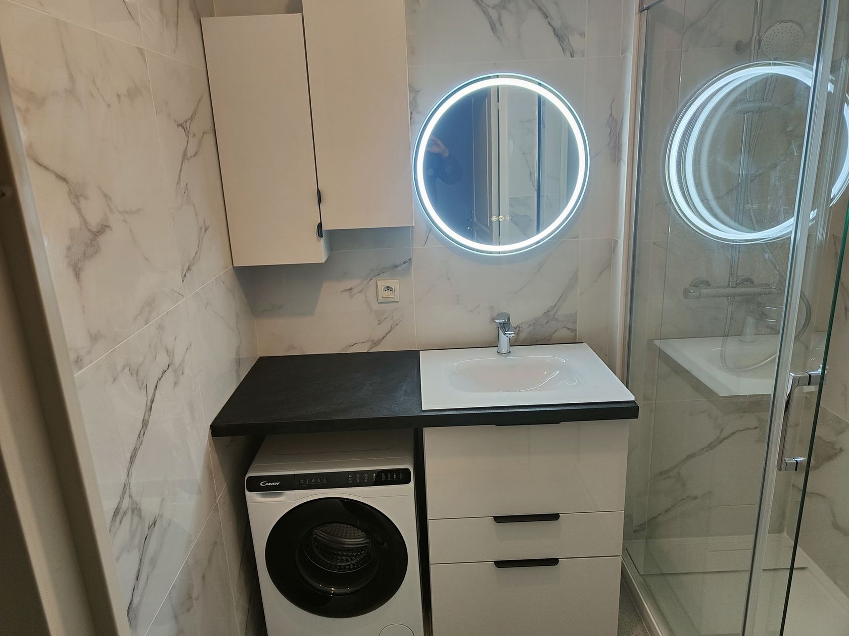 Petite salle de bain avec machine à laver, vanité et miroir circulaire éclairé.