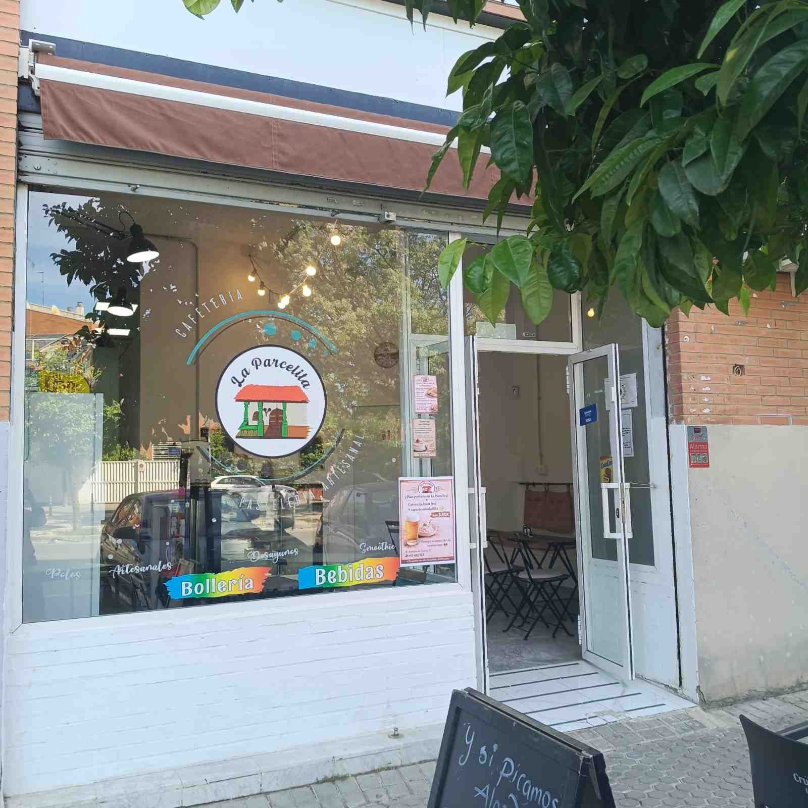 Pequeña tienda con escaparates de cristal, puerta abierta y un árbol en maceta en la acera.