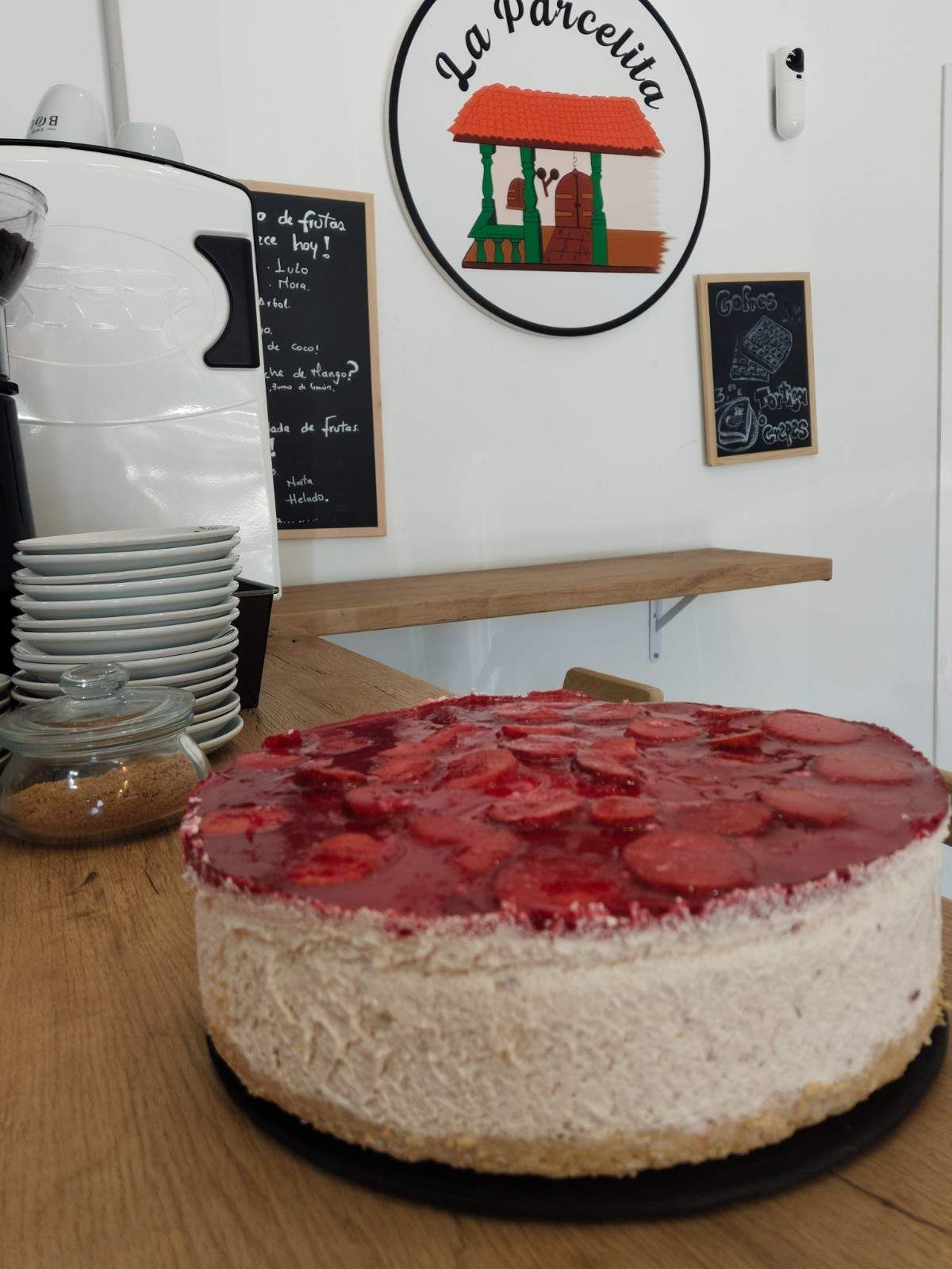 Pastel con fresas sobre un mostrador en una cafetería-panadería con una pizarra y un letrero al fondo.