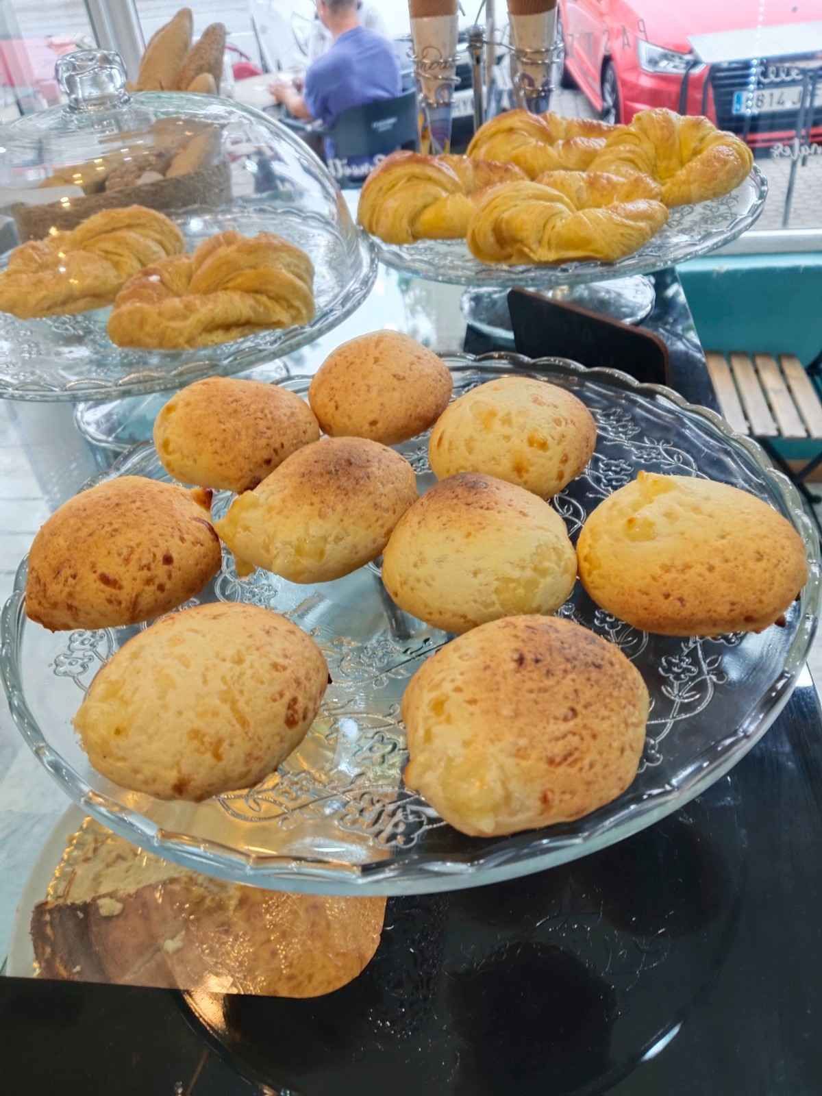 Bandeja de magdalenas redondas y doradas sobre el mostrador de una cafetería, con cruasanes y pan al fondo.