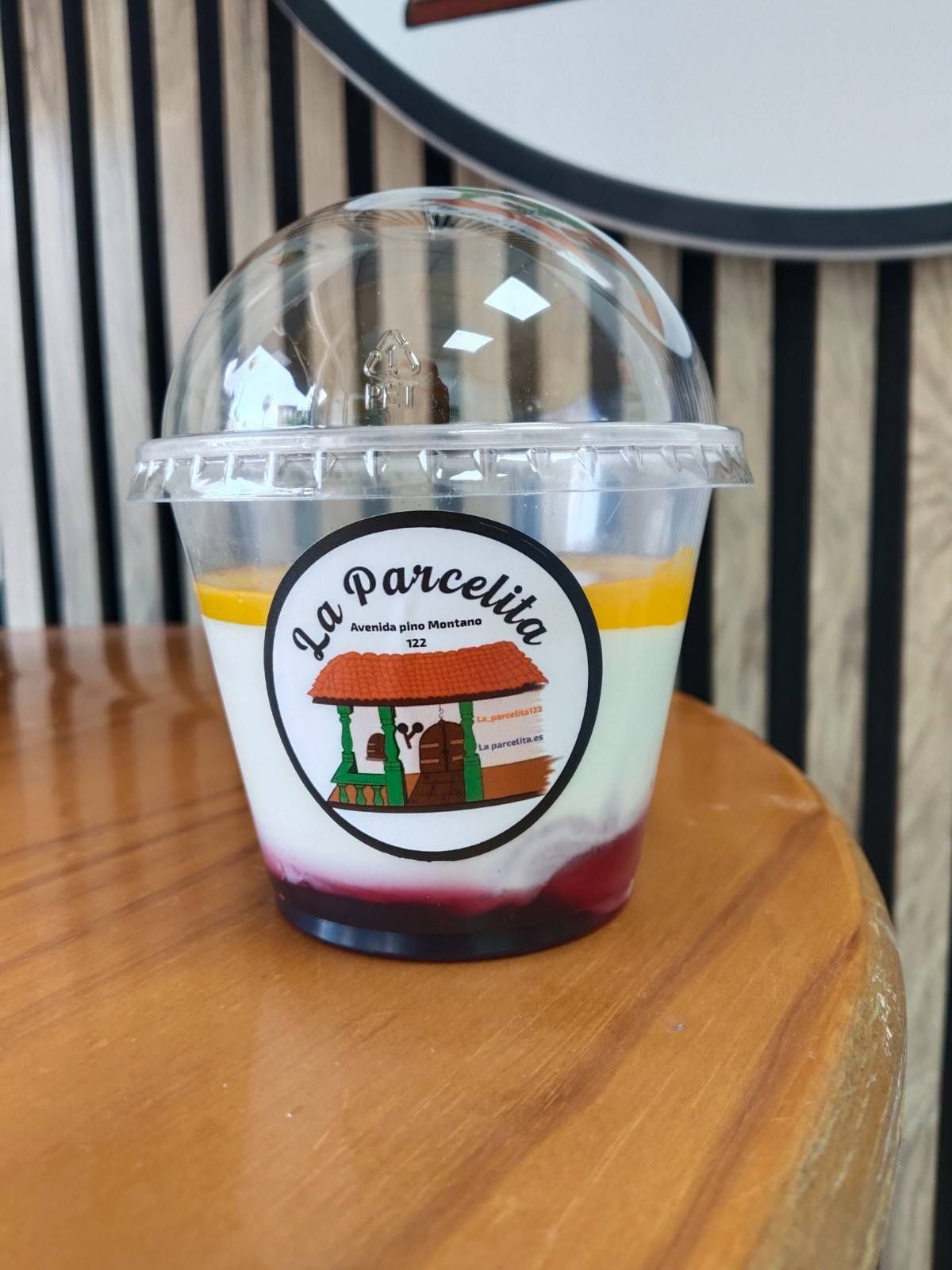 Vaso de postre de plástico transparente con tapa abovedada y cobertura roja sobre una mesa de madera, etiquetado como 