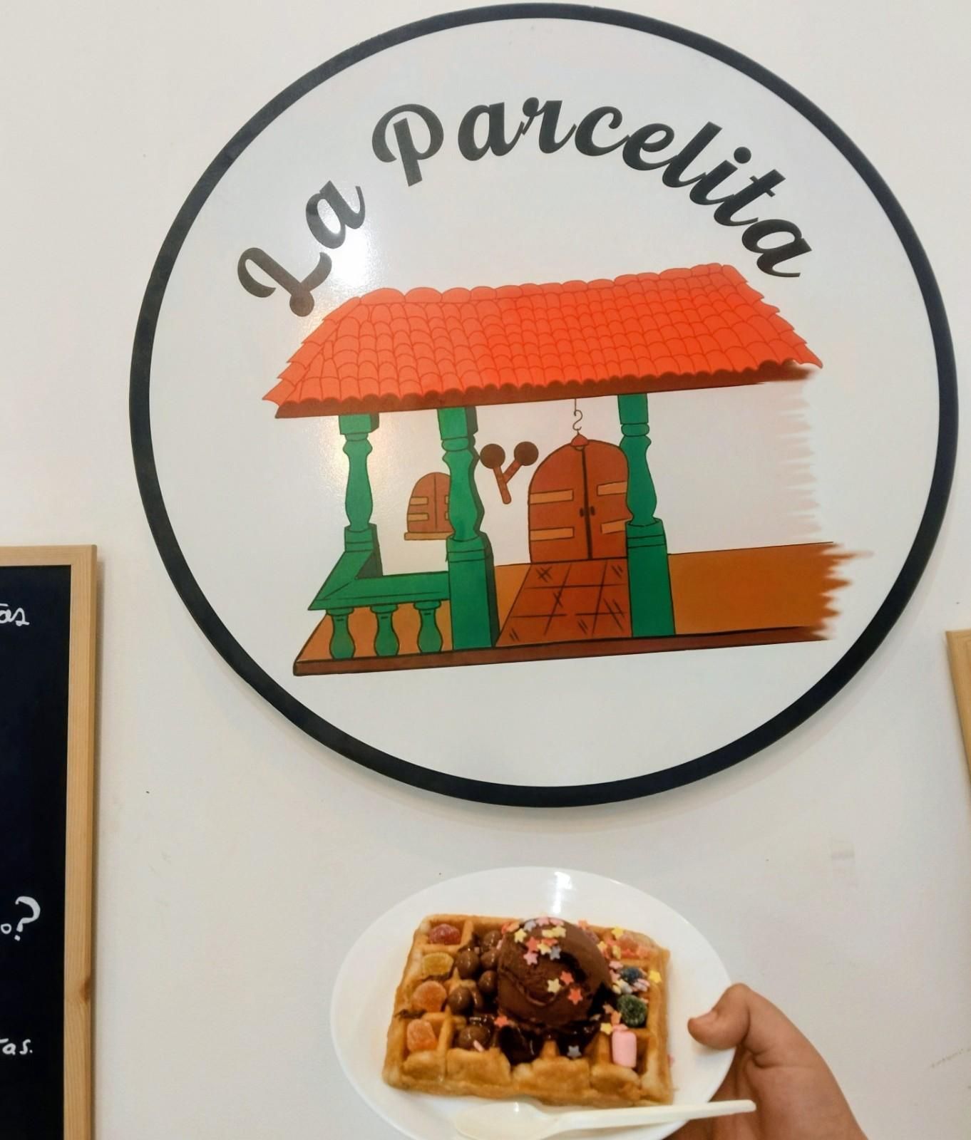 Letrero redondo con el logotipo “La parcelita” con la ilustración de una pequeña casa; debajo, una mano sosteniendo un plato de comida.