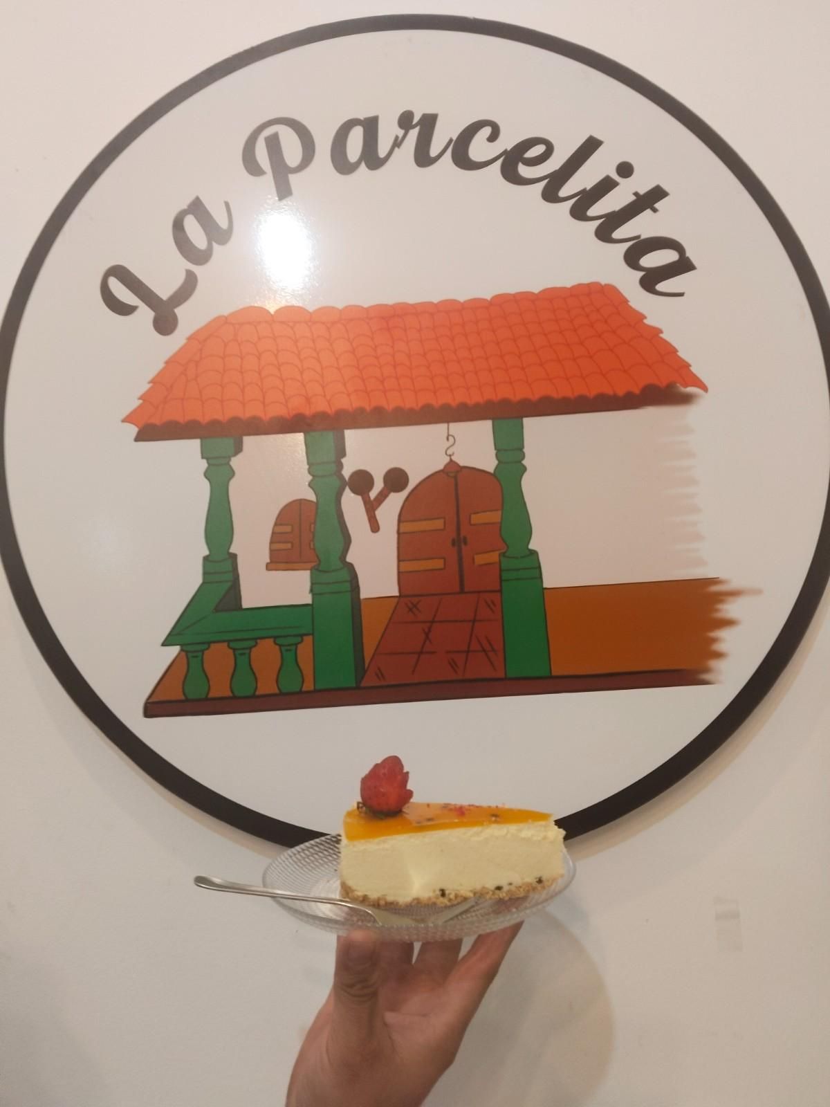 Una mano sostiene una porción de pastel frente a un letrero con el logotipo de “La Parcelita”.