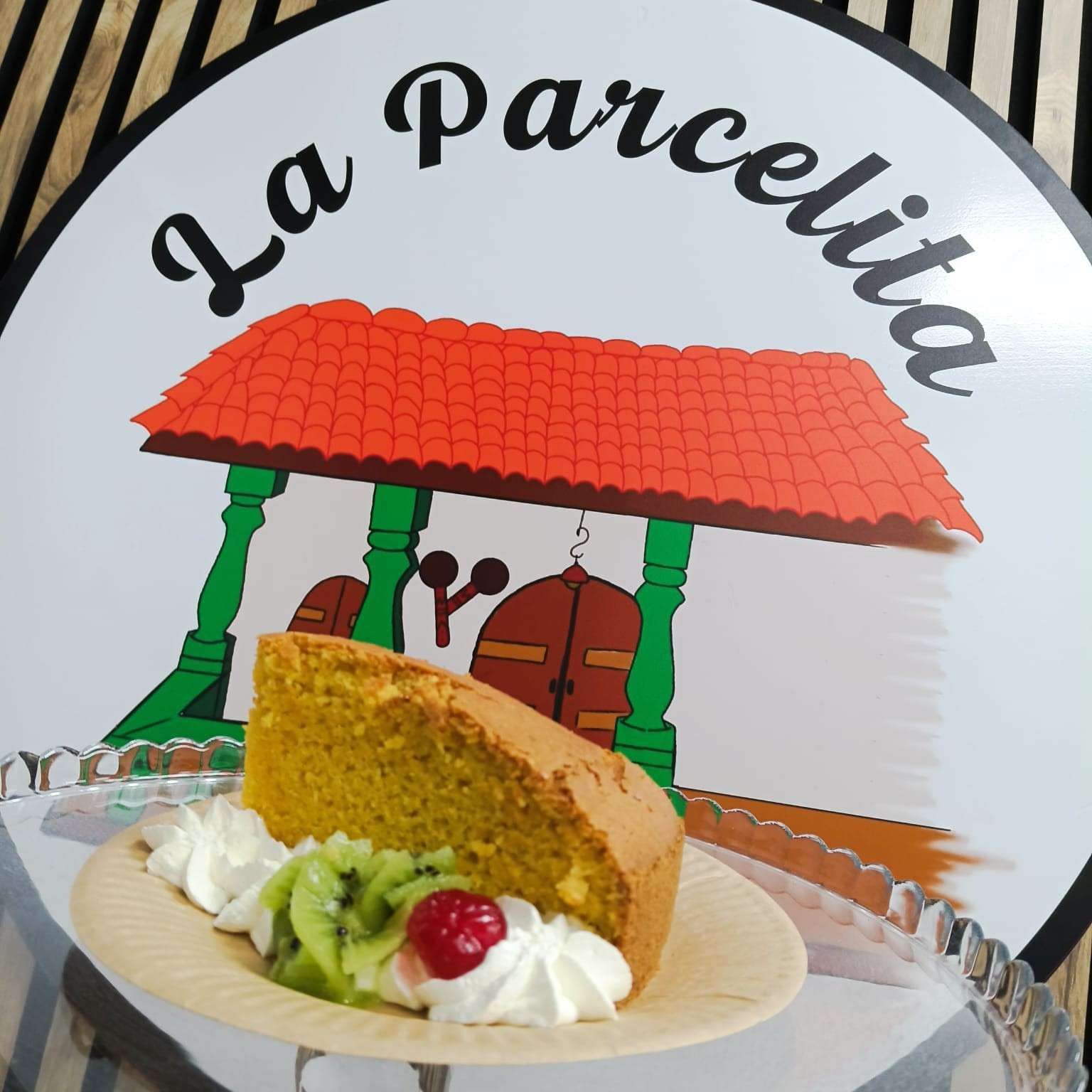 Rebanada de pastel con crema batida y fruta en un plato frente a un letrero de “La Parcellita” y un puesto con techo rojo.