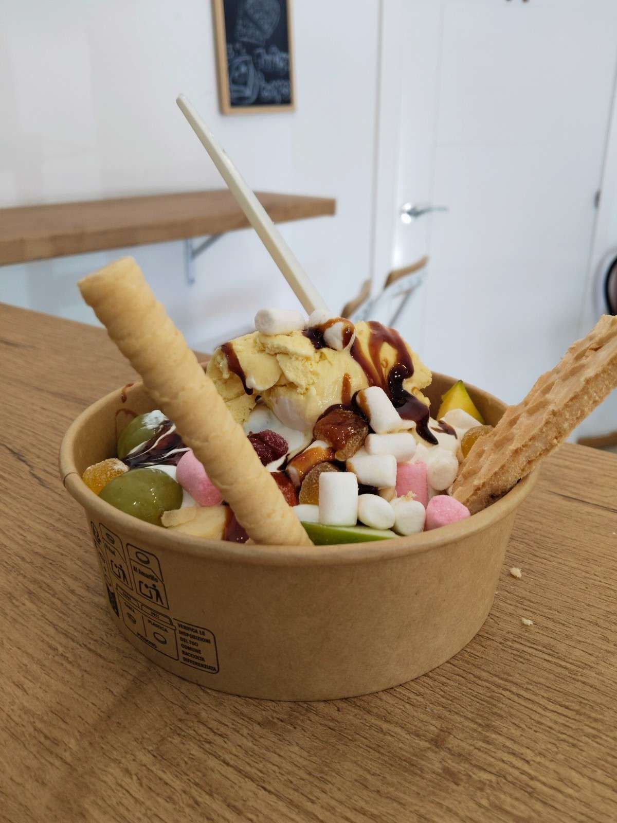 Vaso de yogur helado cubierto con fruta, sirope de chocolate y barquillos sobre una mesa de madera.