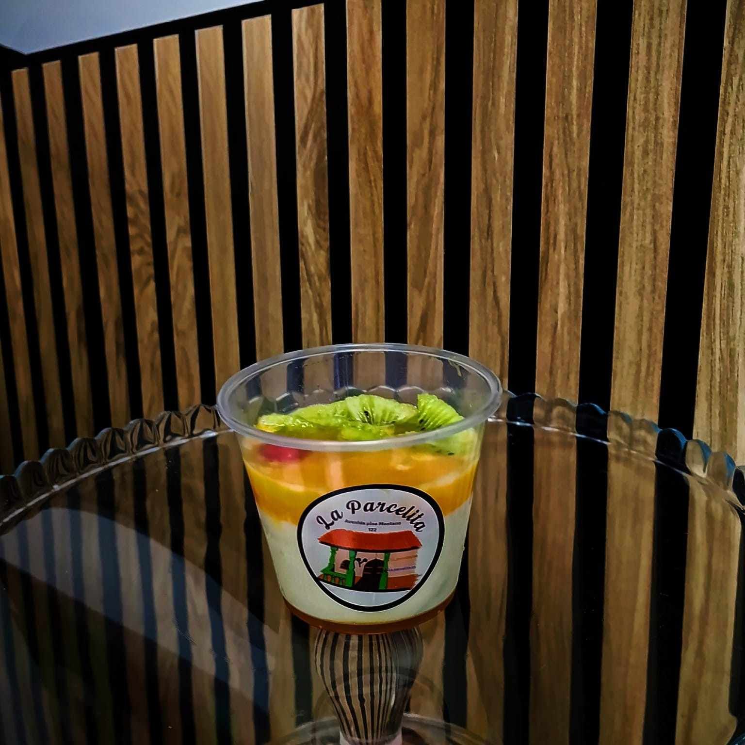 Una taza transparente con postre de frutas y guarnición verde sobre una mesa de cristal con fondo de listones de madera.