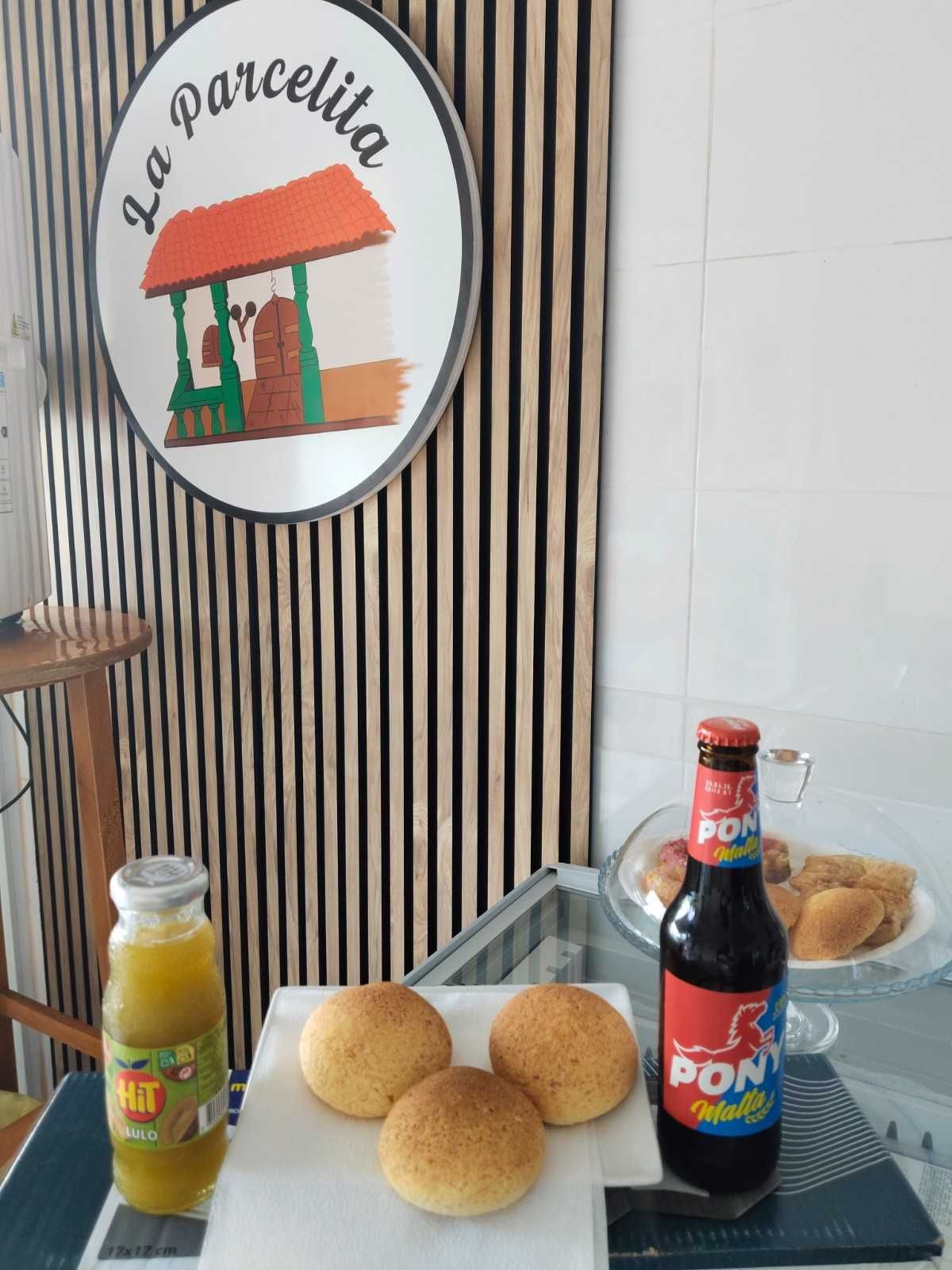 Mostrador de panadería con panecillos, zumo y una botella de refresco junto a un cartel de pared a rayas.