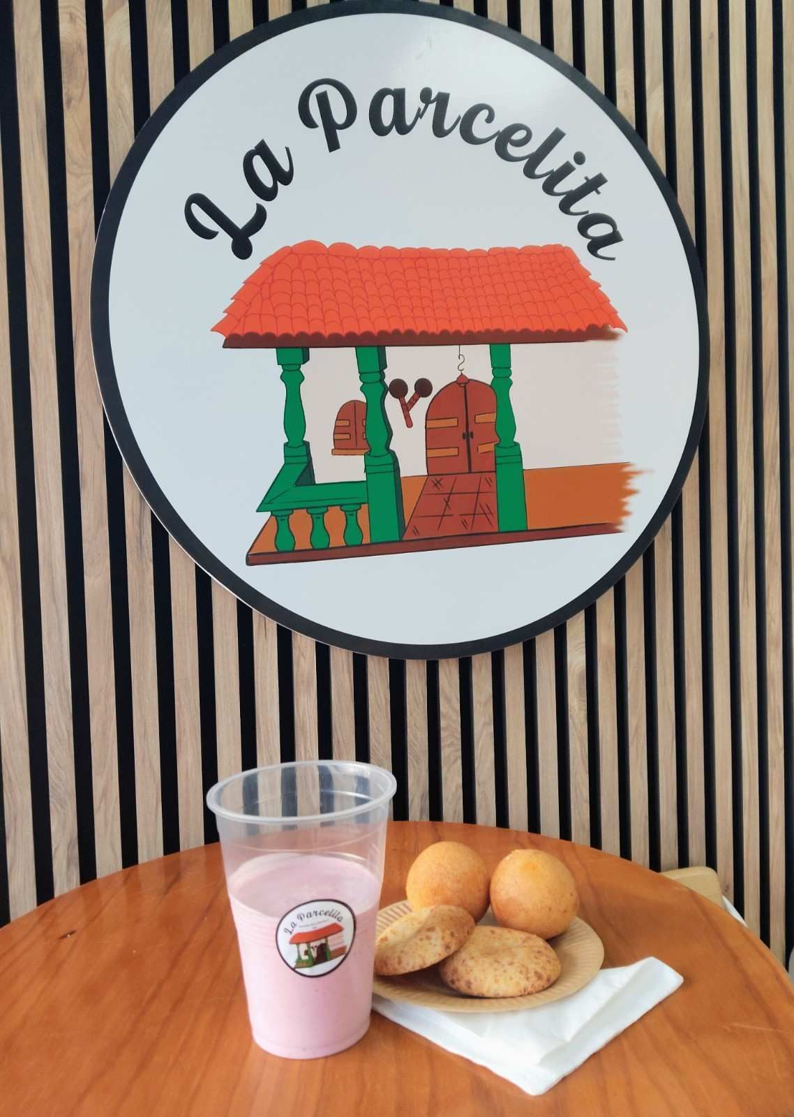 Exhibición de pastelería con un pastel sobre una servilleta y una bebida rosa debajo de un letrero de “La Parcela” y una pared a rayas.