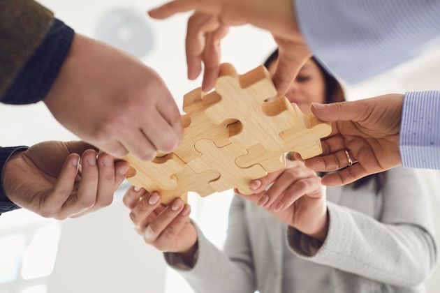 Hände mehrerer Personen, die in einer Teamarbeitsgeste hölzerne Puzzleteile zusammenfügen.