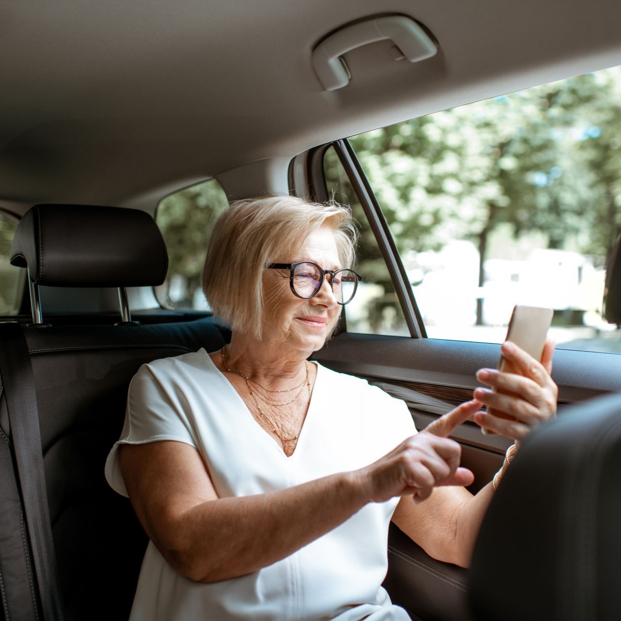 Une femme âgée sur le siège arrière d'une voiture, portant des lunettes, regardant son téléphone.