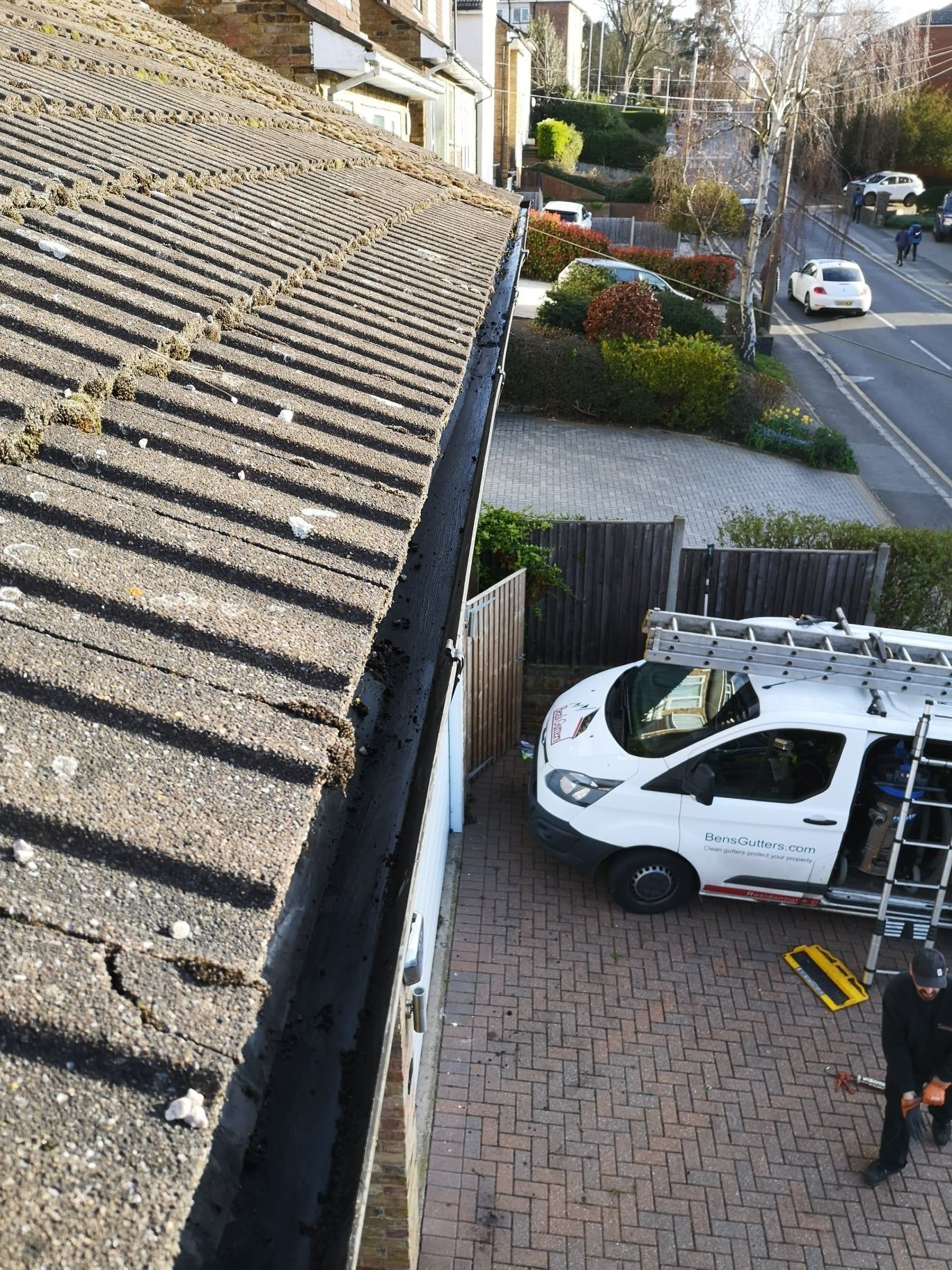 Roofline view of a white service van parked by a house, with a person standing nearby on a brick driveway.