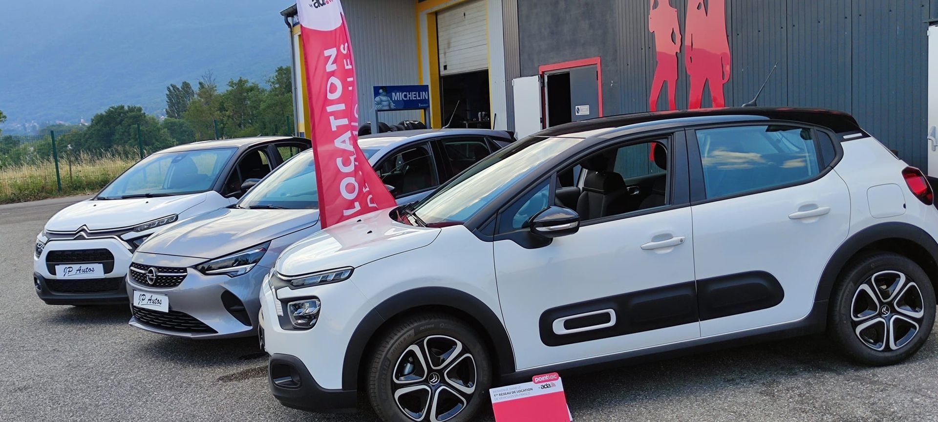 Trois voitures garées devant un garage automobile et voile publicitaire rouge pour promouvoir la location de véhicules