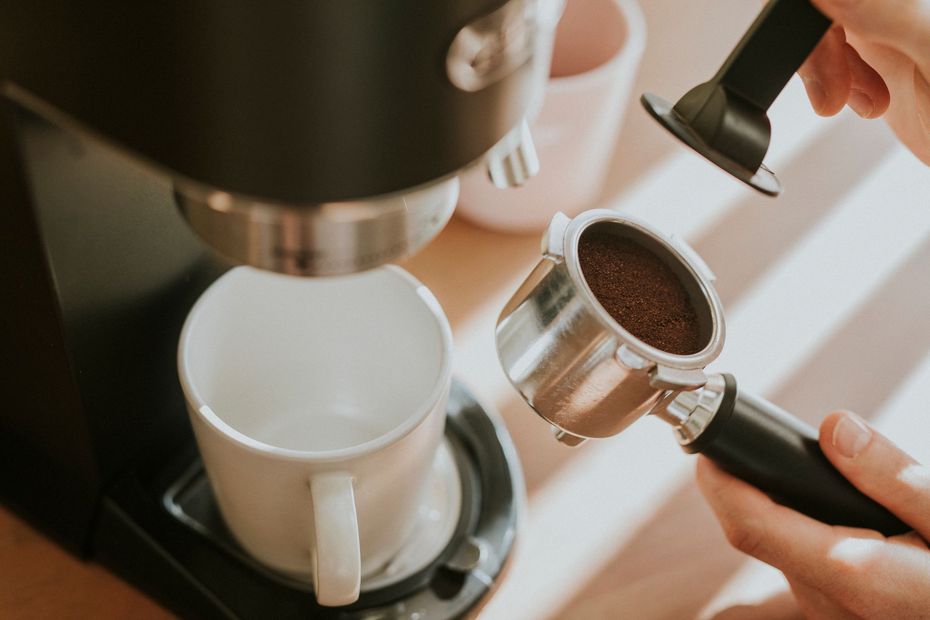 Manos llenando un portafiltro con café molido, junto a una máquina de café y una taza.
