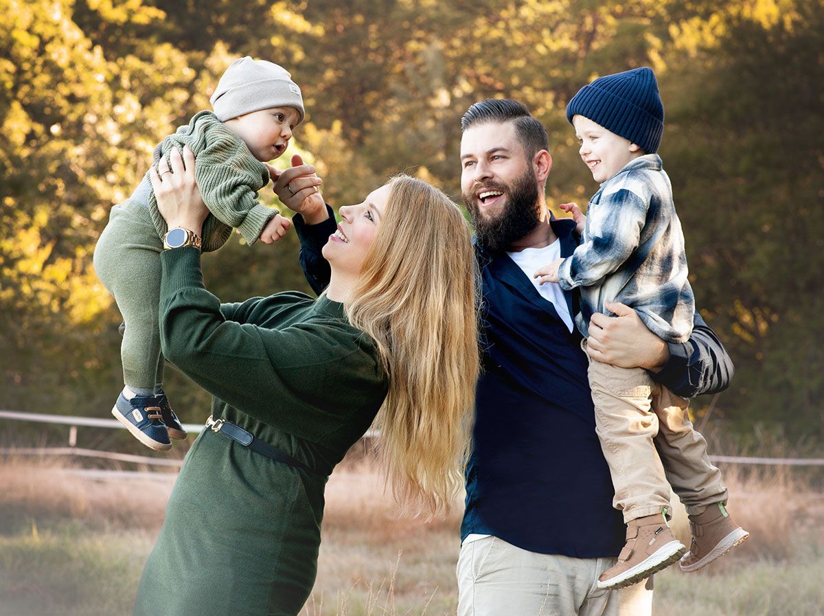 Family of four smiling outdoors: mother holding baby, father holding toddler.