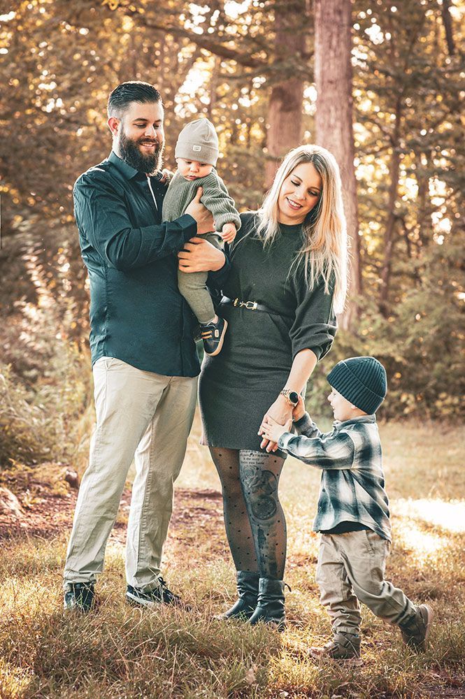 Family of four posing in a forest, mother holding the youngest child, father holding the other, smiles.