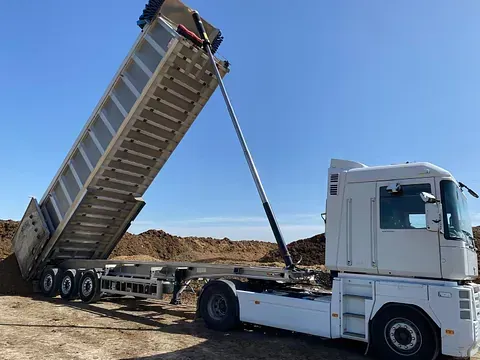 Un camión semirremolque blanco con la caja levantada, arrojando tierra sobre un montón de tierra bajo un cielo azul claro.