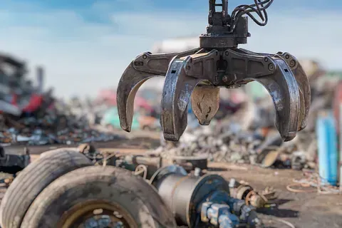 Una grúa con garra de metal sujeta escombros en un depósito de chatarra; en el fondo se ven neumáticos y chatarra.