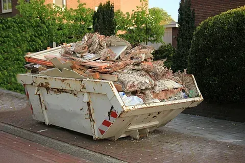Un contenedor de basura blanco repleto de escombros de construcción se encuentra en un área pavimentada al lado de una calle residencial.